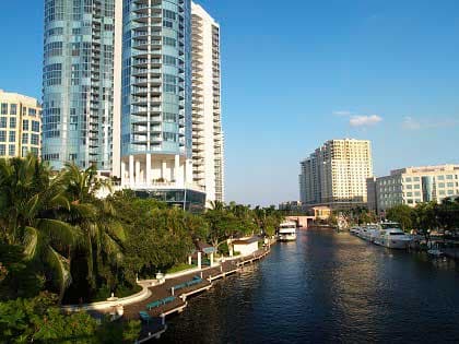 Modern skyscrapers and palm trees line a sunny canal with boats in a city.