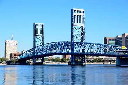 Tall blue drawbridge with two towers crosses river, city buildings in background.